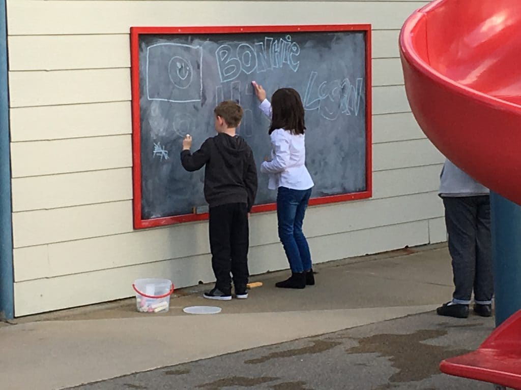 Two children drawing on a chalkboard beside the playground slide